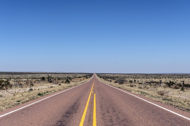 A two-lane road runs straight to the horizon across open high desert under a clear sky.