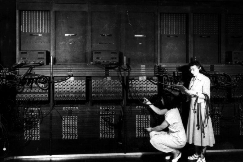 Two operators reprogram the ENIAC computer by rerouting dense bundles of patch cables across its function panels.